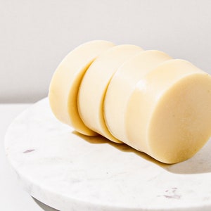 May include: Four round bars of white soap stacked on top of each other on a white marble surface.