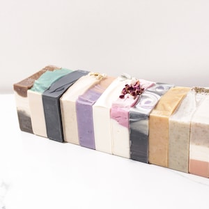May include: A collection of 14 handmade soap bars in various colors and patterns, including green, brown, purple, pink, and white. The bars are arranged in a row, with the ends of the bars visible.