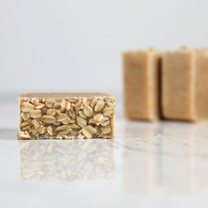 May include: Close-up of a rectangular bar of soap with visible rolled oats embedded on one side. The soap has a light brown color, with three additional bars slightly out of focus in the background. The soap rests on a white surface.