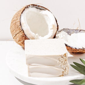 May include: Two bars of white soap with oatmeal on a white marble surface. The soap is next to two halves of a coconut shell.