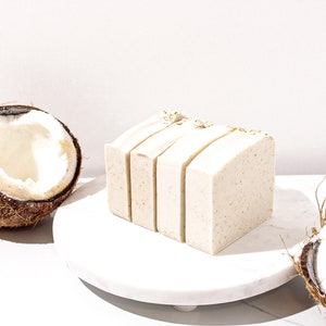 May include: Four bars of white soap with oatmeal and coconut flakes on a white marble surface next to a halved coconut shell.