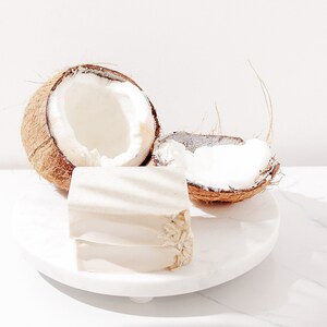 May include: Two halves of a coconut shell with the white flesh exposed, sitting on a white marble surface. A white rectangular bar of soap is in front of the coconut halves.