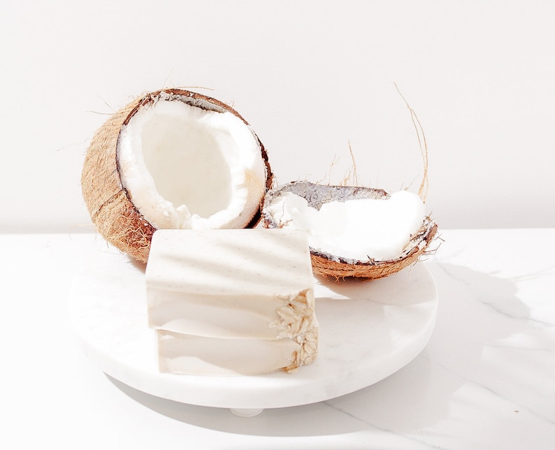 May include: Two halves of a coconut shell with the white flesh exposed, sitting on a white marble surface. A white rectangular bar of soap is in front of the coconut halves.