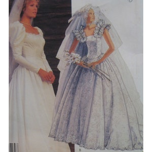 May include: A white wedding dress with long sleeves and a full skirt. The dress has a lace trim and a sweetheart neckline. The model is wearing a veil and holding a bouquet of white flowers.
