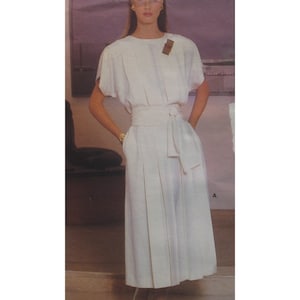 May include: A cream-colored dress with short sleeves, a pleated skirt, and a matching belt. The dress has a simple, elegant design, with a small decorative element near the shoulder. The model is wearing gold earrings and a bracelet.