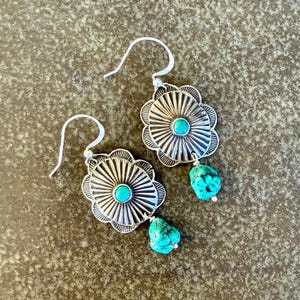 May include: A pair of silver earrings featuring a scalloped, sunburst-patterned metal disc with a small, round turquoise cabochon at the center. A rough, irregular turquoise nugget hangs from the bottom of each silver disc on a hook wire.