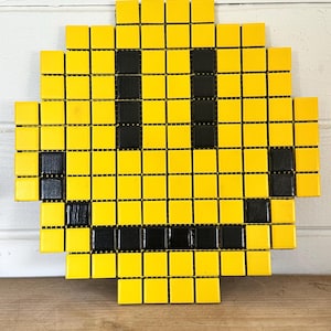May include: A mosaic smiley face made of yellow and black square tiles. The yellow tiles form the face, while the black tiles create the eyes and mouth. The artwork is mounted on a white surface.