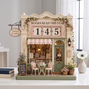 May include: A miniature bookshop model featuring a flip-style counter displaying the number 145 under the text BOOKS READ THIS YEAR. The facade includes a hanging sign reading Emily's Bookshop, a pink striped awning, two chairs, a table, and floral accents.