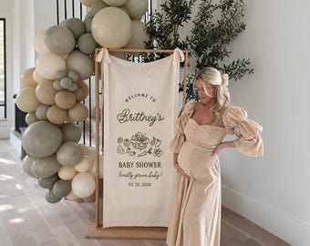 Może przedstawiać: Pionowy, prostokątny baner z tkaniny zawieszony na drewnianej ramie, zawierający napis "WELCOME TO Brittney's BABY SHOWER locally grown baby! 01.10.2026" oraz rysunek liniowy kosza z produktami, ozdobiony dużym łukiem z balonów.