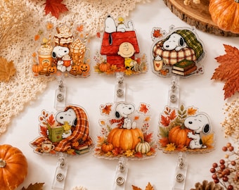 May include: Six acrylic badge reels featuring Snoopy and Charlie Brown in autumn-themed scenes. Designs include Snoopy with pumpkin spice drinks, sitting on a red doghouse, sleeping under a blanket, reading a book, sitting on a pumpkin, and hugging a pumpkin, all surrounded by fall leaves.