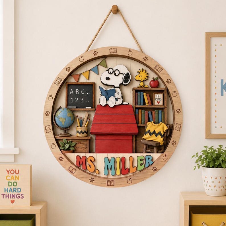 May include: A circular wooden wall sign featuring a 3D scene of Snoopy reading on a red doghouse. The design includes a chalkboard with 'ABC... 123...', a globe, a bookshelf, a yellow shirt, and the name 'MS. MILLER' in colorful 3D letters.