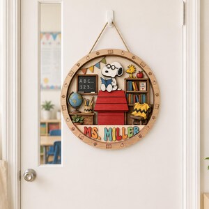 May include: A circular wooden wall sign featuring a 3D Snoopy character reading on a red doghouse, a chalkboard with "ABC... 123...", a bookshelf, a globe, and a yellow shirt. The name "MS. MILLER" is displayed in colorful 3D letters at the bottom.