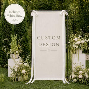 May include: A rectangular off-white fabric banner suspended on a black metal frame. The banner features the text CUSTOM DESIGN in gray serif font with a small heart icon below. Two long white ribbons are tied at the top corners as bows.