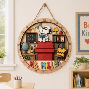May include: A circular wooden wall hanging featuring a 3D scene of Snoopy reading on a red doghouse. The frame is decorated with engraved books, apples, and paw prints. The name MS. MILLER is displayed in colorful 3D letters at the bottom.