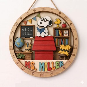 May include: A circular wooden wall hanging featuring a 3D Snoopy character reading a book atop a red doghouse. The scene includes a chalkboard with ABC 123 text, a globe, a bookshelf, a yellow shirt, and the colorful name text MS. MILLER.