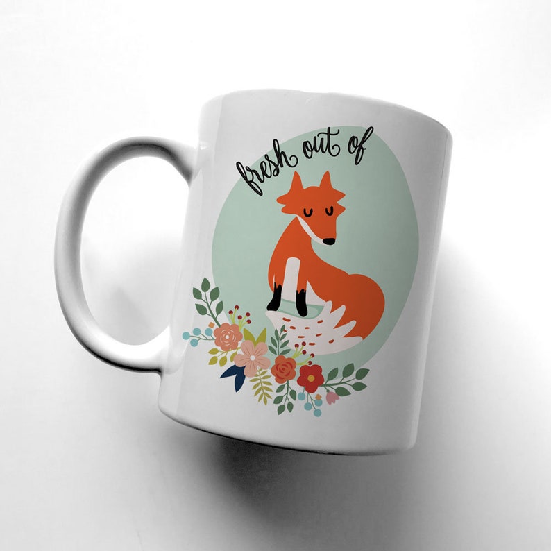May include: White ceramic mug with a fox illustration and the text "fresh out of" in black script. The fox is orange and has a white tail. The background is a light blue circle with a floral design.