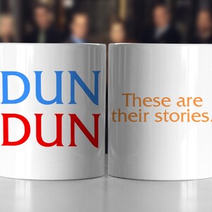 May include: Two white ceramic mugs with a blue and red "DUN DUN" design on one mug and the text "These are their stories." in orange on the other mug.