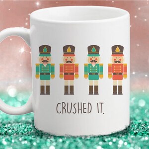 May include: White ceramic mug featuring four colorful nutcracker soldier illustrations. The nutcrackers are in green and red uniforms with black boots and hats. The text "CRUSHED IT." is printed below the illustrations.