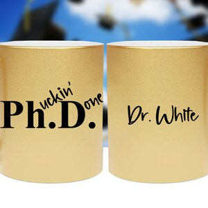 May include: Two gold mugs with black lettering. One mug says "Ph.D.  uckin' one" and the other mug says "Dr. White".