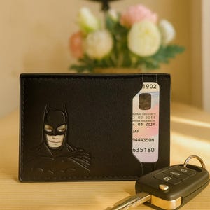 May include: Black leather wallet featuring a Batman graphic. The wallet has a card holder with printed details and is next to a car key. The background includes a decorative globe and flowers.