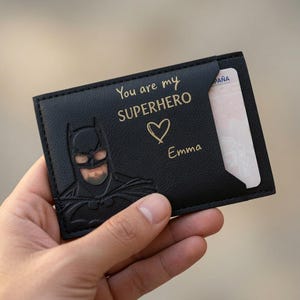 Op de afbeelding: Een zwarte, rechthoekige kaarthouder met een reliëf van een Batman-masker aan de linkerkant. De tekst "You are my SUPERHERO" is in goud gedrukt boven een klein hartje en de naam "Emma" in gouden schrijfletters, met een kaart erin.