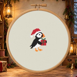 Può includere: Un ricamo a punto croce raffigurante una pulcinella di mare che indossa un cappello da Babbo Natale rosso e bianco, mentre tiene un regalo rosso con un nastro verde. Il disegno è centrato su un tessuto beige chiaro in un telaio circolare.