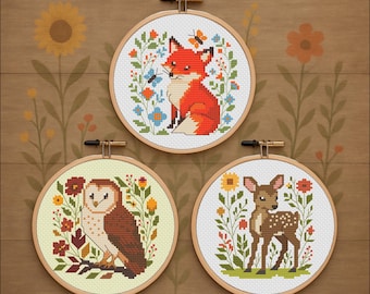 May include: Three circular wooden embroidery hoops containing cross-stitch patterns. The top hoop features a red fox with flowers and a butterfly. The bottom left hoop shows a brown owl on a branch. The bottom right hoop displays a spotted fawn.