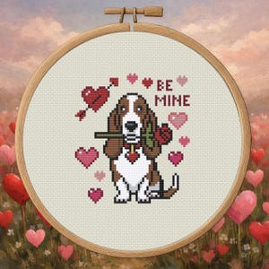 Könnte beinhalten: Ein Kreuzstich-Stickrahmen mit einem Basset Hound, der eine rote Rose im Maul hält. Das Design zeigt rosa Herzen, einen Pfeil durch ein Herz und die Worte "BE MINE". Der Rahmen ist vor einem Hintergrund aus Herzen angeordnet.