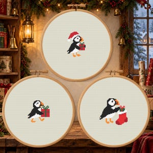 May include: Three circular wooden embroidery hoops containing cross-stitch patterns on off-white fabric. Each hoop features a black and white puffin with an orange beak. One puffin wears a Santa hat and holds a gift, another carries a gift, and the third holds a red Christmas stocking.