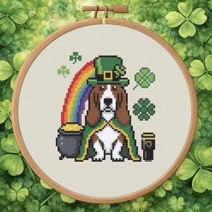 May include: Cross-stitch embroidery in a wooden hoop features a dog dressed as a leprechaun. The dog wears a green hat and cape, with a rainbow, pot of gold, and shamrocks. The design is on a light beige fabric.