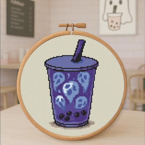 May include: Cross-stitch embroidery in a wooden hoop depicts a purple bubble tea cup with ghost designs. The cup has a lid and a straw. The background is a neutral beige fabric. A small framed picture of a ghost holding bubble tea is in the background.