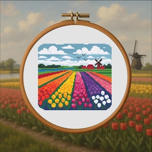 May include: Cross-stitch embroidery of a colorful tulip field with a windmill and red-roofed buildings under a blue sky with clouds. The embroidery is set in a wooden hoop, showcasing a vibrant landscape.