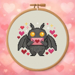 Könnte beinhalten: Kreuzstich-Stickerei in einem Holzreifen. Das Design zeigt eine graue Fledermaus, die einen rosa Umschlag mit einem Herz hält, umgeben von rosa und roten Herzen und gelben und weißen Glitzer. Die Fledermaus hat gelbe Augen.