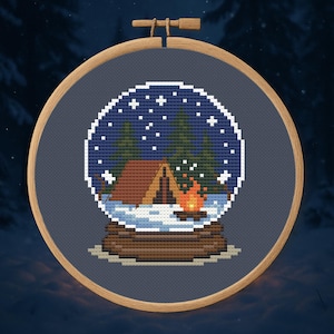 Puede incluir: Bordado de punto de cruz en un aro de madera. El diseño presenta una escena de globo de nieve con una tienda de campaña, una fogata y pinos bajo un cielo estrellado. El aro mide aproximadamente 18 cm de diámetro.