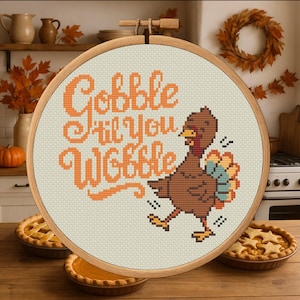 May include: A cross-stitch design in a wooden hoop features the phrase "Gobble Til You Wobble" in orange letters. A cartoon turkey with brown feathers and a colorful tail is also included. The background shows pies and autumn decor.