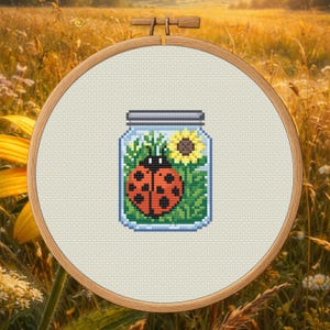 May include: A cross-stitch embroidery hoop featuring a jar design. Inside the jar is a ladybug with red and black spots, a yellow sunflower, and green foliage. The hoop is set against a blurred background of a field.