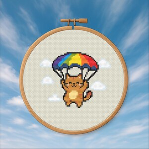 May include: Cross-stitch embroidery in a wooden hoop features a pixelated orange cat with a rainbow parachute. The cat is smiling, and the background includes white clouds against a blue sky. The embroidery is a craft item.