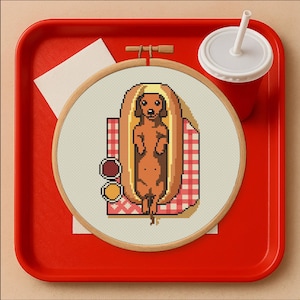 May include: A cross-stitch embroidery hoop showing a dachshund dog in a hot dog bun, on a red and white checked picnic blanket. The embroidery is on cream fabric, with condiment circles. A red tray and drink complete the scene.