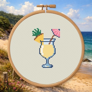 Può includere: Ricamo a punto croce in un telaio di legno raffigurante un cocktail pixelato con una fetta di ananas e un ombrellino rosa. La bevanda è gialla, su un tessuto neutro. Lo sfondo mostra una scena di spiaggia con acqua e cielo blu.