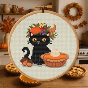 Könnte beinhalten: Kreuzstich-Stickerei in einem Holzrahmen mit einer schwarzen Katze, die einen Herbstkranz trägt und neben einem Kürbiskuchen sitzt. Zwei Eicheln liegen zu Füßen der Katze. Der Hintergrund umfasst Herbstdekorationen und Kuchen.