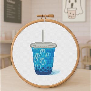 Puede incluir: Un diseño de punto de cruz de una bebida de té de burbujas azul con cubitos de hielo con forma de fantasma, exhibido en un aro de bordado de madera. La bebida tiene una tapa gris y una pajita. El fondo incluye una ilustración de fantasma y un menú.