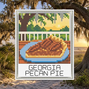 May include: A cross-stitch pattern featuring a slice of pecan pie on a blue plate, set on a wooden table with a white railing and trees in the background. The text GEORGIA PECAN PIE is printed in black at the bottom of the rectangular design.