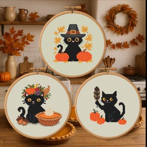 Könnte beinhalten: Drei runde Kreuzstich-Stickrahmen mit schwarzen Katzen mit Halloween- und Herbstmotiven. Eine Katze trägt einen Hexenhut, eine andere einen Blumenkranz, und die dritte hält eine Feder. Kürbisse und Herbstblätter sind ebenfalls enthalten.
