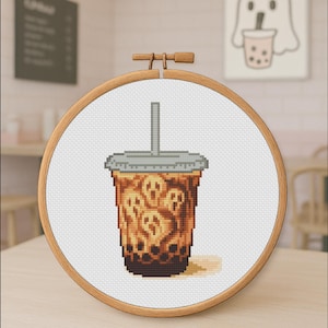 May include: Cross-stitch embroidery of a bubble tea drink with ghost faces. The drink is in a clear plastic cup with a grey lid and straw. The embroidery is set in a wooden hoop, with a light-coloured background.