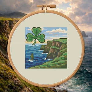 Puede incluir: Bordado de punto de cruz de un trébol verde sobre un paisaje costero con acantilados y océano. La obra de arte está enmarcada en un bastidor de bordado de madera, sobre un fondo de acantilados y cielo nublado.