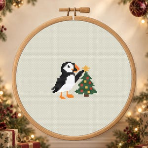 Puede incluir: Un patrón de punto de cruz en un bastidor de madera redondo que muestra un frailecillo blanco y negro con patas naranjas colocando una estrella amarilla sobre un árbol de Navidad verde decorado con pequeños adornos rojos y amarillos sobre tela crema.