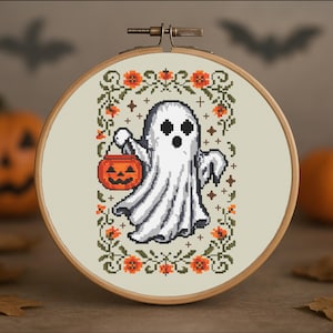 May include: A cross-stitch embroidery in a wooden hoop features a white ghost holding an orange jack-o'-lantern bucket. The ghost is surrounded by a floral border with orange flowers and green leaves, perfect for Halloween decor.
