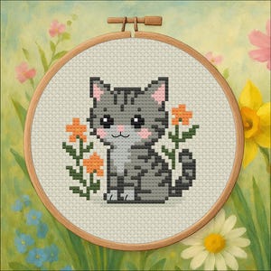 Puede incluir: Un bordado de punto de cruz de un gato atigrado gris con orejas y mejillas rosas, rodeado de flores naranjas. El gato está centrado dentro de un aro de bordado de madera, sobre un fondo de flores y vegetación pintadas.