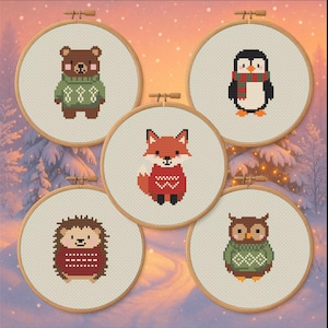 Op de afbeelding: Vijf borduurringen met kruissteek met bosdieren: een beer, een pinguïn, een vos, een egel en een uil. Elk dier draagt een trui. De ringen staan tegen een besneeuwde, winterse achtergrond.