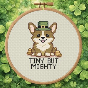 May include: Cross-stitch embroidery of a corgi wearing a green leprechaun hat, surrounded by gold coins. The text "TINY BUT MIGHTY" is stitched below the corgi. The embroidery is set in a wooden hoop, against a background of green shamrocks.
